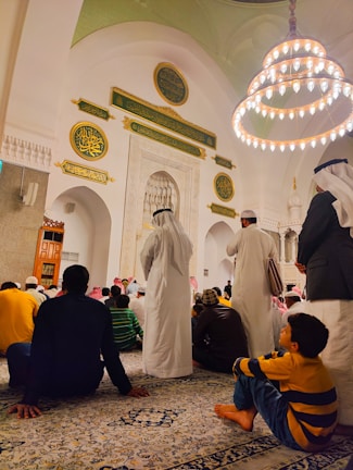A gathering of people inside a mosque, with intricate Islamic architecture featuring Arabic calligraphy and a large chandelier hanging from the ceiling. Many of the attendees are sitting on a patterned carpet. Some are dressed in traditional Middle Eastern attire, such as white robes and keffiyehs.