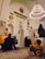 A gathering of people inside a mosque, with intricate Islamic architecture featuring Arabic calligraphy and a large chandelier hanging from the ceiling. Many of the attendees are sitting on a patterned carpet. Some are dressed in traditional Middle Eastern attire, such as white robes and keffiyehs.