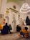 A gathering of people inside a mosque, with intricate Islamic architecture featuring Arabic calligraphy and a large chandelier hanging from the ceiling. Many of the attendees are sitting on a patterned carpet. Some are dressed in traditional Middle Eastern attire, such as white robes and keffiyehs.