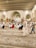 A large indoor space with ornate architectural details and a patterned carpet is filled with people sitting or walking. The ceiling features large, elegant arches and hanging chandeliers. The crowd appears diverse and engaged in a communal activity, possibly prayer or a gathering.