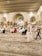 A large indoor space with ornate architectural details and a patterned carpet is filled with people sitting or walking. The ceiling features large, elegant arches and hanging chandeliers. The crowd appears diverse and engaged in a communal activity, possibly prayer or a gathering.