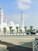 A large, white mosque featuring multiple domes and minarets stands under a clear blue sky. Palm trees line the area in front of the mosque, and a police car is parked on the street nearby.