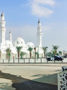 A large, white mosque featuring multiple domes and minarets stands under a clear blue sky. Palm trees line the area in front of the mosque, and a police car is parked on the street nearby.
