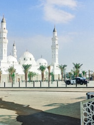 A large, white mosque featuring multiple domes and minarets stands under a clear blue sky. Palm trees line the area in front of the mosque, and a police car is parked on the street nearby.