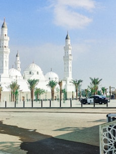 A large, white mosque featuring multiple domes and minarets stands under a clear blue sky. Palm trees line the area in front of the mosque, and a police car is parked on the street nearby.