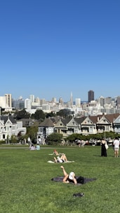 A green park area with several people relaxing on the grass. In the background, a row of Victorian-style houses can be seen, with a city skyline featuring skyscrapers under a clear blue sky.