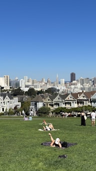 A green park area with several people relaxing on the grass. In the background, a row of Victorian-style houses can be seen, with a city skyline featuring skyscrapers under a clear blue sky.
