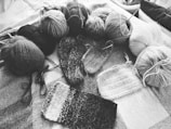 Various balls of yarn are arranged on a textured surface alongside several swatches of knitted fabric. The image is in black and white, emphasizing the textures and patterns of the yarn and knitting. Knitting needles are also visible, indicating the ongoing knitting process.