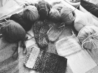 Various balls of yarn are arranged on a textured surface alongside several swatches of knitted fabric. The image is in black and white, emphasizing the textures and patterns of the yarn and knitting. Knitting needles are also visible, indicating the ongoing knitting process.