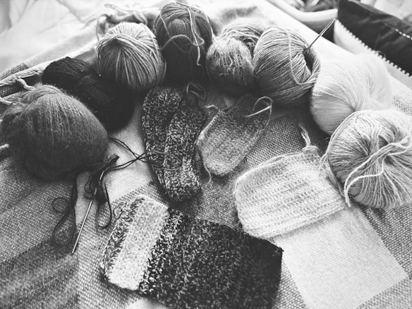 Various balls of yarn are arranged on a textured surface alongside several swatches of knitted fabric. The image is in black and white, emphasizing the textures and patterns of the yarn and knitting. Knitting needles are also visible, indicating the ongoing knitting process.