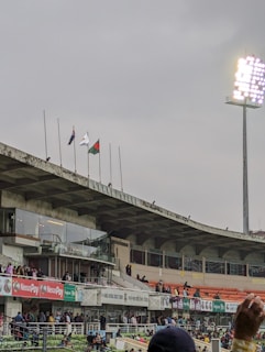 An outdoor stadium scene featuring stands with seated and standing spectators. Flags are hoisted above the seating area, and a tall floodlight tower is prominently lit. Advertising banners and signs are placed around the stadium. The sky appears overcast, suggesting cloudy weather.