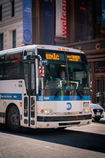 A New York City bus with the route number 'SIM1C' displayed on the front, moving down a city street. The bus is white with blue accents and has the MTA logo. Surrounding the bus are urban buildings with glass facades and banners.