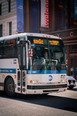 A New York City bus with the route number 'SIM1C' displayed on the front, moving down a city street. The bus is white with blue accents and has the MTA logo. Surrounding the bus are urban buildings with glass facades and banners.