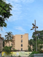 An apartment building surrounded by greenery and palm trees is situated under a clear blue sky. A streetlight with multiple lamps is visible, as well as electrical wires overhead. A sign with text is displayed in front of the building, partially obscured by a white fence.