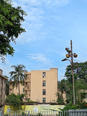 An apartment building surrounded by greenery and palm trees is situated under a clear blue sky. A streetlight with multiple lamps is visible, as well as electrical wires overhead. A sign with text is displayed in front of the building, partially obscured by a white fence.