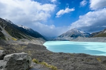 A stunning landscape featuring a turquoise glacial lake surrounded by rocky terrain and towering snow-capped mountains under a dramatic cloudy sky.