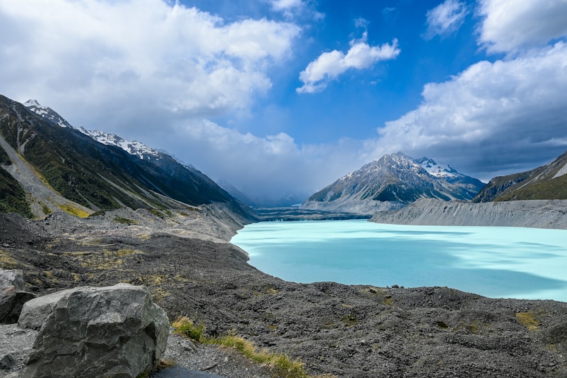 A stunning landscape featuring a turquoise glacial lake surrounded by rocky terrain and towering snow-capped mountains under a dramatic cloudy sky.