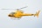 A bright yellow helicopter is flying in the sky, showcasing sleek aerodynamic design with visible registration marks. The helicopter has a single main rotor and a tail rotor.