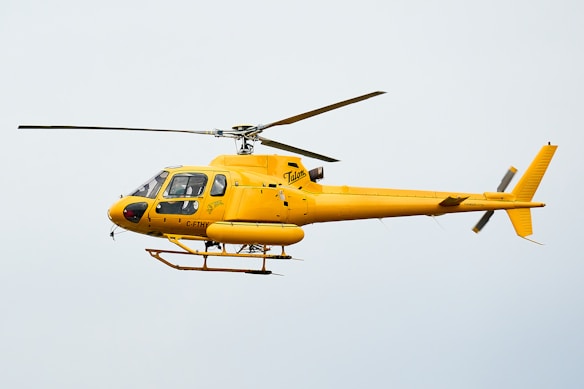 A bright yellow helicopter is flying in the sky, showcasing sleek aerodynamic design with visible registration marks. The helicopter has a single main rotor and a tail rotor.