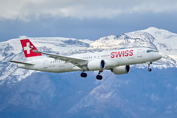 A white commercial airplane with 'SWISS' written in red on the fuselage flies in front of a backdrop of snow-capped mountains. The Swiss flag is visible on the tail fin of the aircraft, and the landing gear is extended as the plane approaches for landing.