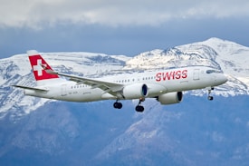 A white commercial airplane with 'SWISS' written in red on the fuselage flies in front of a backdrop of snow-capped mountains. The Swiss flag is visible on the tail fin of the aircraft, and the landing gear is extended as the plane approaches for landing.