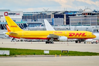 A DHL cargo airplane is parked on the tarmac of an airport with a large building and terminal in the background. The plane is painted in bright yellow and red with the slogan 'AS ONE AGAINST CANCER 2022' along its side. Several other vehicles and small equipment are present on the ground around the aircraft.