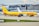 A DHL cargo airplane is parked on the tarmac of an airport with a large building and terminal in the background. The plane is painted in bright yellow and red with the slogan 'AS ONE AGAINST CANCER 2022' along its side. Several other vehicles and small equipment are present on the ground around the aircraft.