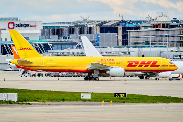 A DHL cargo airplane is parked on the tarmac of an airport with a large building and terminal in the background. The plane is painted in bright yellow and red with the slogan 'AS ONE AGAINST CANCER 2022' along its side. Several other vehicles and small equipment are present on the ground around the aircraft.