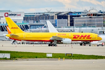 A DHL cargo airplane is parked on the tarmac of an airport with a large building and terminal in the background. The plane is painted in bright yellow and red with the slogan 'AS ONE AGAINST CANCER 2022' along its side. Several other vehicles and small equipment are present on the ground around the aircraft.