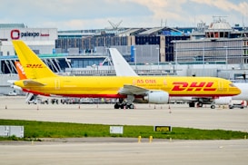 A DHL cargo airplane is parked on the tarmac of an airport with a large building and terminal in the background. The plane is painted in bright yellow and red with the slogan 'AS ONE AGAINST CANCER 2022' along its side. Several other vehicles and small equipment are present on the ground around the aircraft.