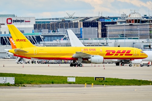 A DHL cargo airplane is parked on the tarmac of an airport with a large building and terminal in the background. The plane is painted in bright yellow and red with the slogan 'AS ONE AGAINST CANCER 2022' along its side. Several other vehicles and small equipment are present on the ground around the aircraft.