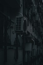 A row of air conditioning units mounted on an exterior wall of a building with a dark and moody lighting. The units are lined up in a repetitive pattern alongside decorative metalwork and a tiled wall.