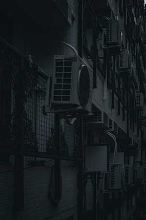 A row of air conditioning units mounted on an exterior wall of a building with a dark and moody lighting. The units are lined up in a repetitive pattern alongside decorative metalwork and a tiled wall.