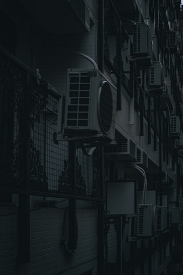 A row of air conditioning units mounted on an exterior wall of a building with a dark and moody lighting. The units are lined up in a repetitive pattern alongside decorative metalwork and a tiled wall.