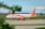 A passenger airplane with the easyJet logo is parked on a runway. The aircraft is predominantly white with orange accents, including orange engines and a striking orange tail featuring the airline's branding. The background shows a grassy field, with some distant trees and a blue sky with light clouds visible. The foreground features blurred leaves, adding a touch of greenery to the scene.