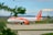 A passenger airplane with the easyJet logo is parked on a runway. The aircraft is predominantly white with orange accents, including orange engines and a striking orange tail featuring the airline's branding. The background shows a grassy field, with some distant trees and a blue sky with light clouds visible. The foreground features blurred leaves, adding a touch of greenery to the scene.