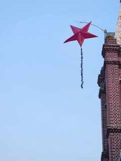 A red star-shaped kite is entangled on the upper corner of a brick building. The sky is clear and blue. The kite has a long tail with black adornments.