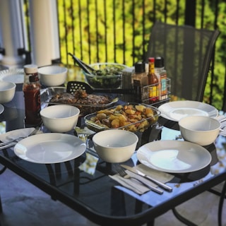 A glass dining table is set outdoors, surrounded by chairs. It features white plates and bowls, folded napkins, and cutlery arranged neatly. In the center, there are dishes including baked potatoes and a salad, with various sauces and dressings in bottles nearby. Greenery in the background suggests a garden or balcony setting.
