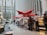 A modern indoor cafe with bright red triangular umbrellas suspended above the counter. The cafe is located in a spacious atrium with tall glass windows and concrete pillars. Several people are standing in line, interacting with the staff at the counter, which displays an assortment of food items. Comfortable seating is arranged with round tables in the foreground.