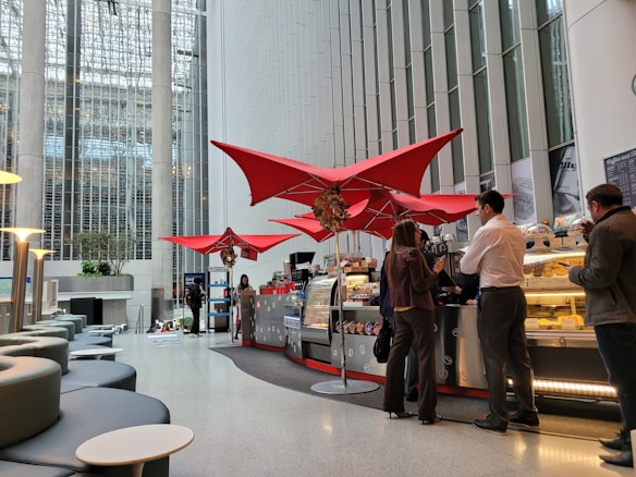 A modern indoor cafe with bright red triangular umbrellas suspended above the counter. The cafe is located in a spacious atrium with tall glass windows and concrete pillars. Several people are standing in line, interacting with the staff at the counter, which displays an assortment of food items. Comfortable seating is arranged with round tables in the foreground.
