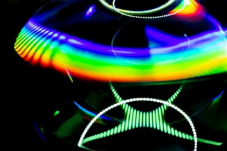A vibrant display of abstract light patterns featuring a spectrum of colors primarily in a rainbow gradient. The image contains circular and wavy lines with a luminous and reflective quality, set against a dark background that enhances the vividness of the colors.