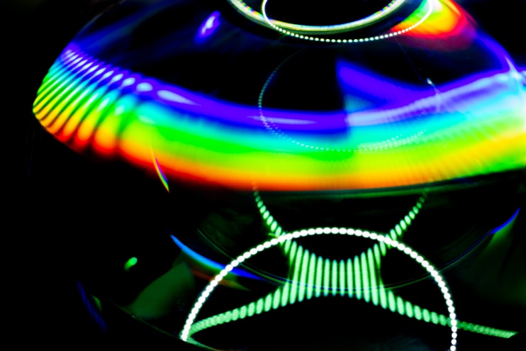 A vibrant display of abstract light patterns featuring a spectrum of colors primarily in a rainbow gradient. The image contains circular and wavy lines with a luminous and reflective quality, set against a dark background that enhances the vividness of the colors.