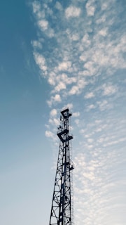 A tall communication tower stands prominently against a sky filled with scattered, fluffy clouds. The structure is composed of lattice-like metal work, rising vertically with various platforms and equipment attached. The overall scene suggests an interplay between technology and nature.