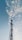 A tall communication tower stands prominently against a sky filled with scattered, fluffy clouds. The structure is composed of lattice-like metal work, rising vertically with various platforms and equipment attached. The overall scene suggests an interplay between technology and nature.