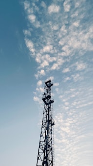 A tall communication tower stands prominently against a sky filled with scattered, fluffy clouds. The structure is composed of lattice-like metal work, rising vertically with various platforms and equipment attached. The overall scene suggests an interplay between technology and nature.