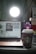 A cozy workspace with a laptop displaying an out-of-focus screen, a smartphone showing the time, and a branded coffee cup in the foreground. A bright light source illuminates the scene from behind the laptop.