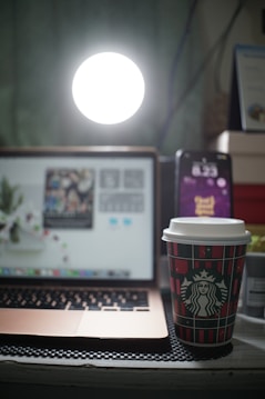 A cozy workspace with a laptop displaying an out-of-focus screen, a smartphone showing the time, and a branded coffee cup in the foreground. A bright light source illuminates the scene from behind the laptop.