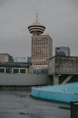 A cityscape featuring a prominent tower with a unique UFO-like design on top, surrounded by other modern buildings. The foreground shows a curved concrete structure with a light blue railing. The sky is overcast, casting a muted light over the scene.