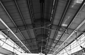 An interior view of a large industrial or commercial building featuring a vast ceiling with exposed metal beams and a corrugated metal roof. There are rows of evenly spaced windows that allow natural light to filter through, casting shadows on the structure.