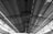 An interior view of a large industrial or commercial building featuring a vast ceiling with exposed metal beams and a corrugated metal roof. There are rows of evenly spaced windows that allow natural light to filter through, casting shadows on the structure.